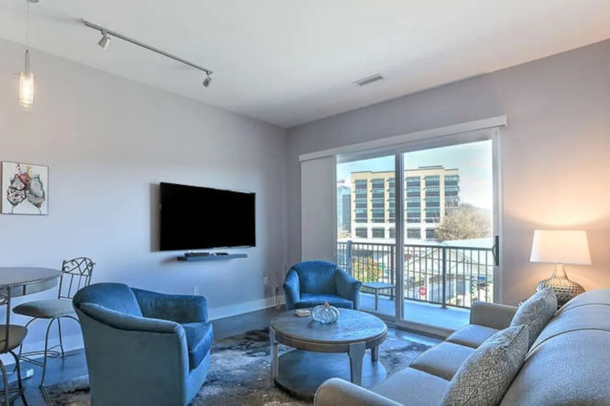 A bright and spacious living room featuring a large window that frames a scenic city view, with a comfortable sofa, blue armchairs, a modern coffee table adorned with decorative pieces, and a wall-mounted TV. Soft lighting creates a cozy atmosphere, and balcony access is visible, connecting the interior to the vibrant city outside.