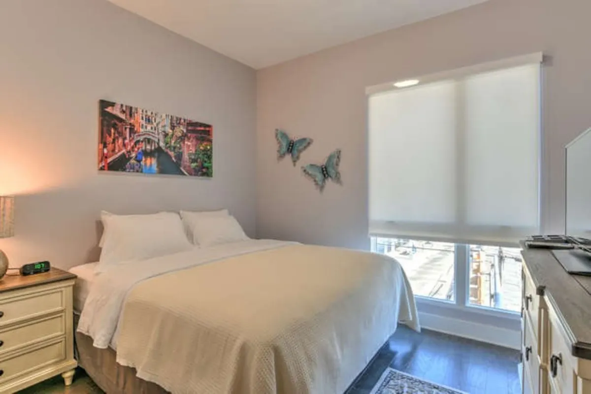A welcoming king bedroom featuring a plush bed adorned with luxurious bedding, soft lighting from a nightstand lamp, and soothing butterfly artwork above the headboard. A painting of a tranquil canal scene adds an artistic touch, with natural morning light filtering through the windows.