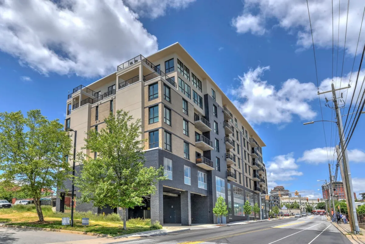 The modern exterior of the apartment building is animated by balconies overlooking the city, green trees lining the sidewalk, and a vibrant, lively atmosphere. The building's facade establishes a direct visual connection between the interior lounge and downtown Asheville.